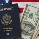 A United States passport lies next to a stack of hundred-dollar bills on top of an American flag, symbolizing the intersection of immigration policy and financial remittances.