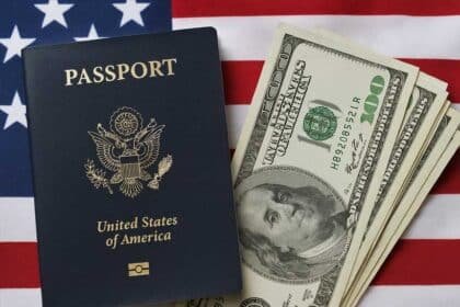 A United States passport lies next to a stack of hundred-dollar bills on top of an American flag, symbolizing the intersection of immigration policy and financial remittances.